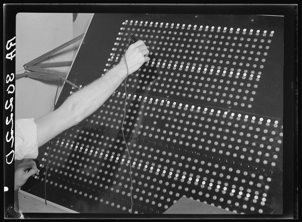 A hand wiring a control panel of many rows of sockets with a patch cord, an early plugboard used to program a machine by physical configuration